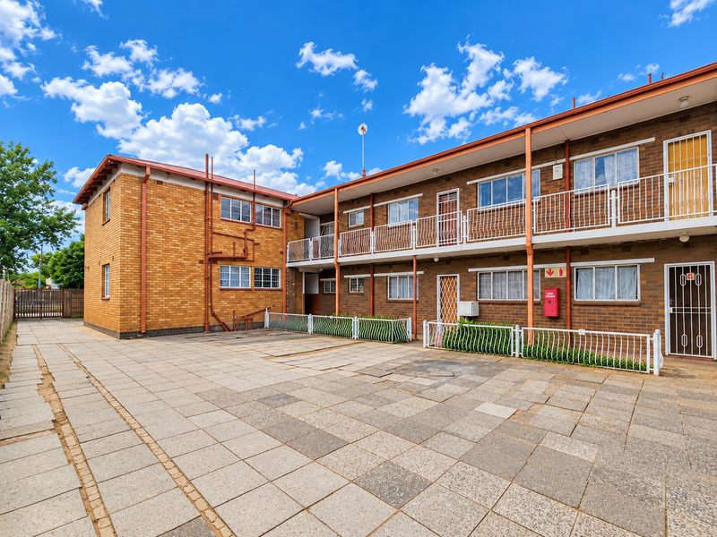 Rear view of a brick apartment building featuring a paved courtyard, metal balcony railings, and security gates on the ground floor units.