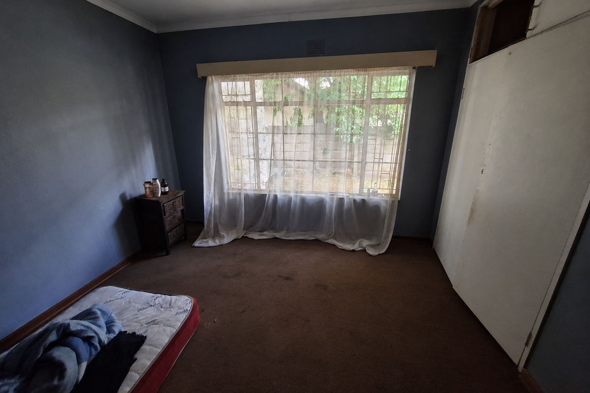 A bedroom featuring a large window, built-in wardrobe with missing panel, and carpet flooring. The room requires cosmetic updates to improve its overall condition.