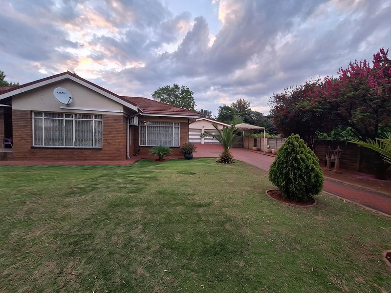 A well-maintained single-story brick home featuring a spacious front lawn, paved driveway, double garage, and carport under a dramatic sunset sky.