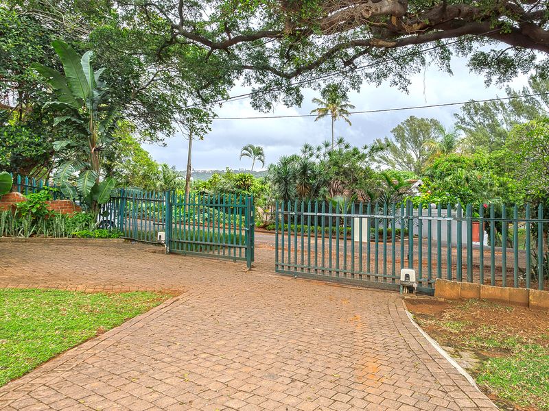 A view of the property entrance featuring a paved driveway, mature trees, and a secure metal gate leading to the street.