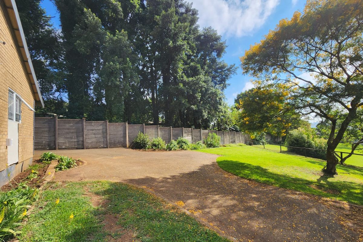 A spacious rear garden featuring a paved driveway, established lawn, mature trees, and a concrete boundary fence.
