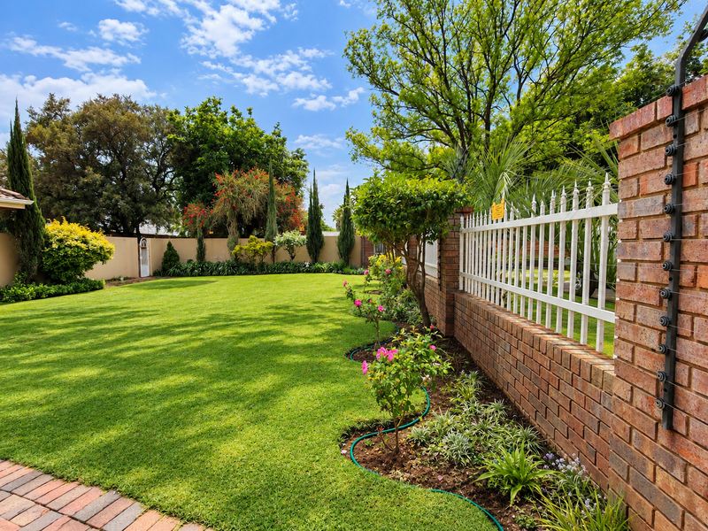 A well-maintained garden featuring a lush green lawn, landscaped flower beds, a brick boundary wall with electric fencing, and a paved walkway.