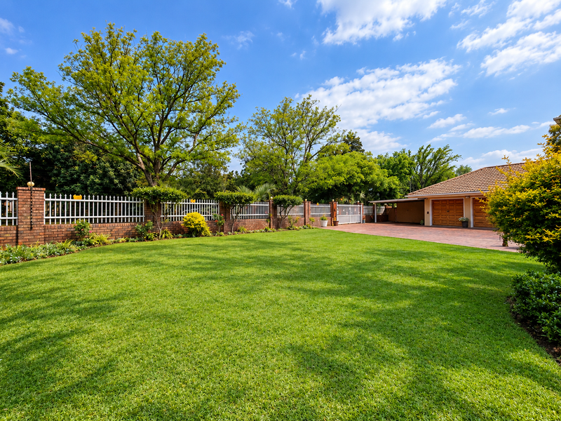 A well-maintained front garden featuring a lush green lawn, brick boundary wall with metal fencing, and a paved driveway leading to a double garage.