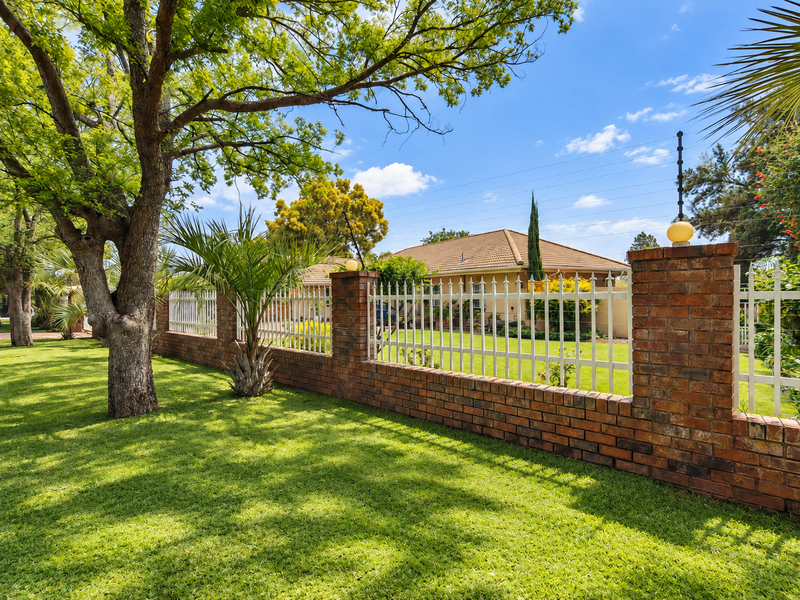 A well-maintained garden featuring a brick and metal boundary wall with integrated electric fencing, mature trees, and a lush green lawn.