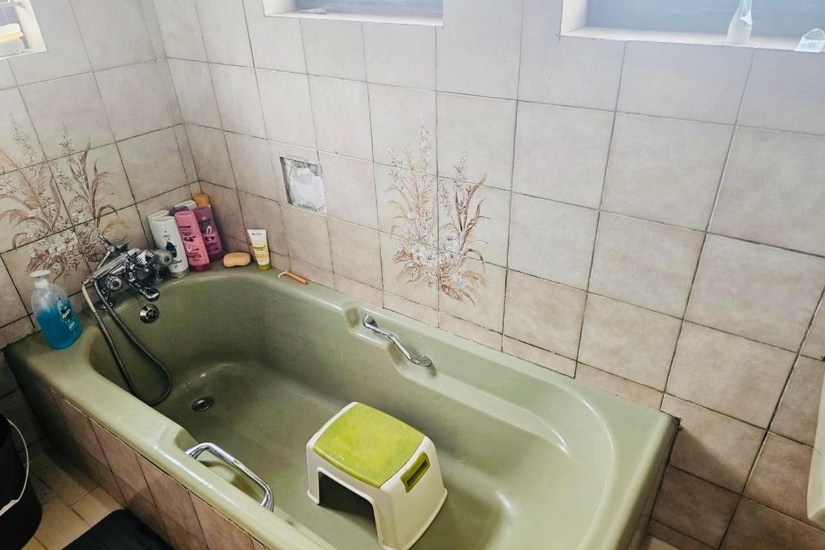 A dated bathroom featuring a green bathtub with integrated mixer tap and handheld shower, finished with decorative ceramic wall tiles.