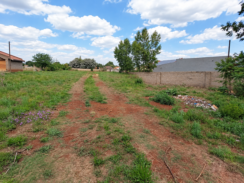 A vacant residential plot featuring an unpaved access track, perimeter brick walling, and established grass coverage.