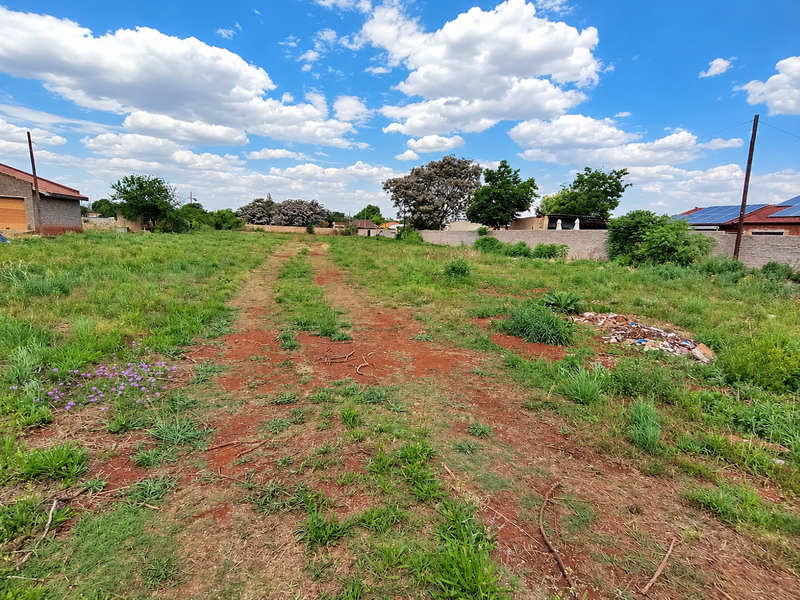 A vacant residential plot featuring natural grass, red soil, and boundary walls, offering a clear space for future development.