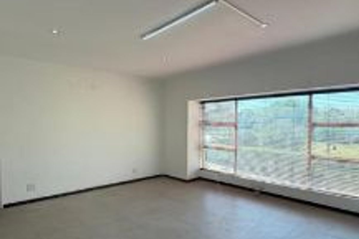 A bright, empty living room featuring large windows with blinds, neutral tiled flooring, and a combination of recessed and surface-mounted lighting.