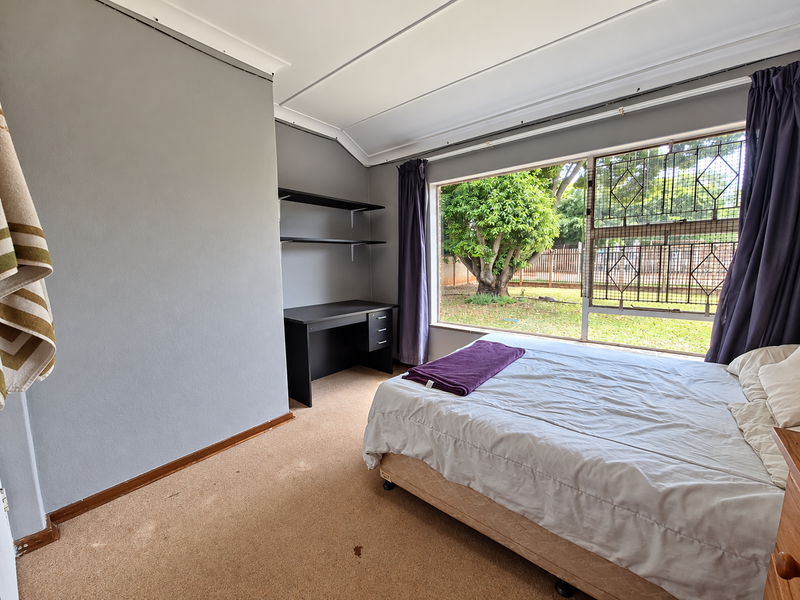 A bedroom featuring carpeted flooring, built-in shelving, and a large window with security bars overlooking the garden.