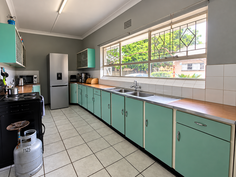A functional kitchen featuring retro-style cabinetry, a double stainless steel sink, tiled splashbacks, and large windows with security bars.