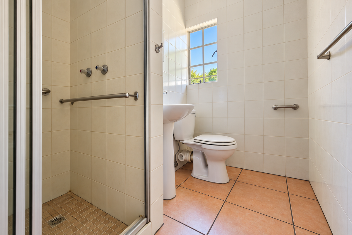 A functional bathroom featuring a walk-in shower, pedestal basin, toilet, and safety grab rails, finished with neutral wall tiles and terracotta floor tiles.