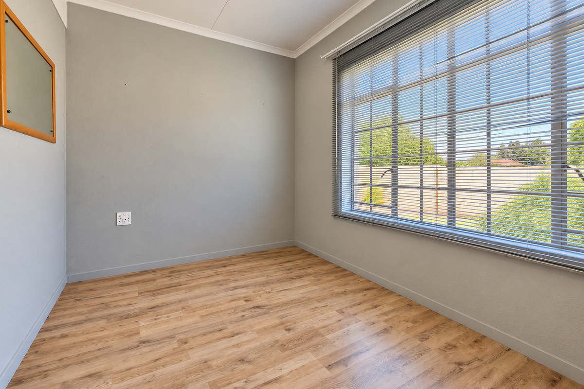 A bright and neutral empty bedroom featuring modern laminate flooring, recessed lighting, and large windows fitted with venetian blinds.