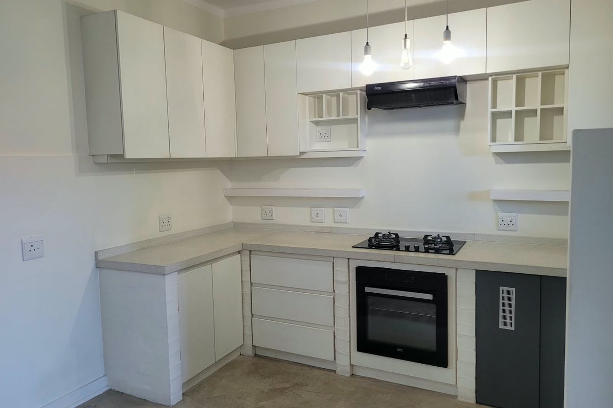 A clean, modern kitchen featuring white built-in cabinetry, stone countertops, a built-in oven, gas hob, and extractor fan with minimalist pendant lighting.