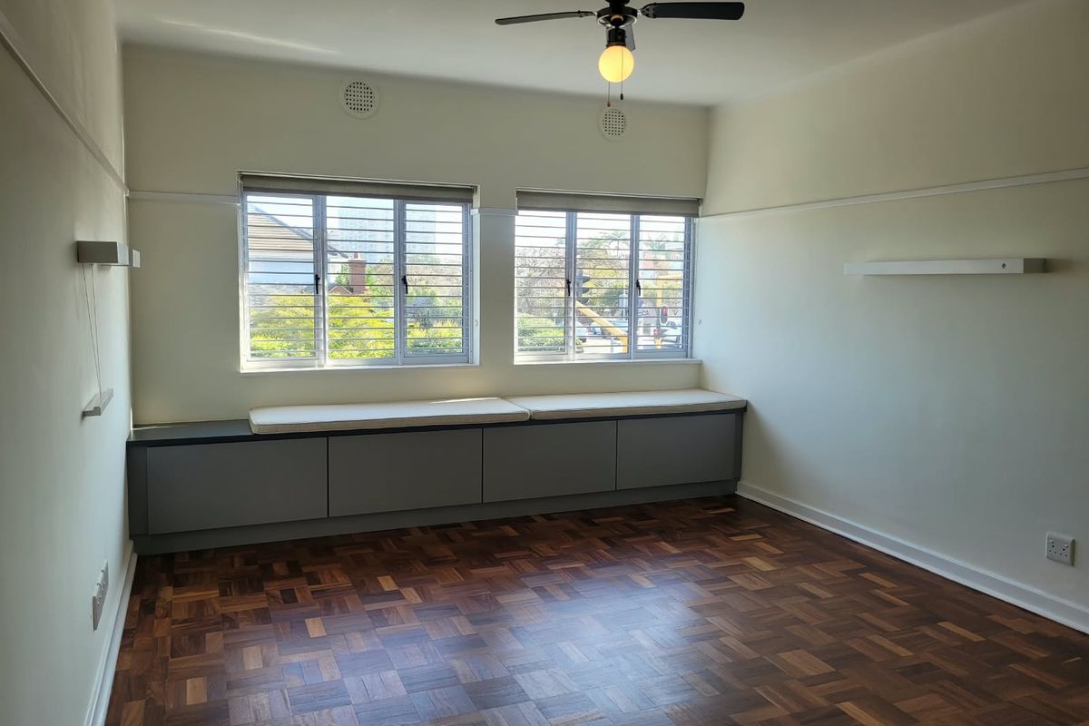 A bright, empty bedroom featuring classic parquet flooring, a ceiling fan, and custom built-in window seat cabinetry with integrated storage.