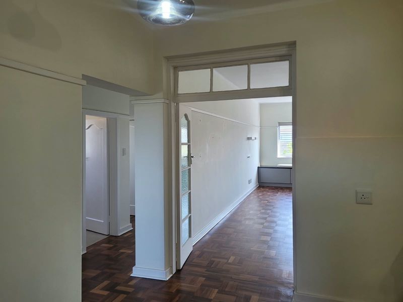A bright, empty hallway featuring classic parquet flooring, a glass-paneled door with a transom window, and traditional picture rail detailing.