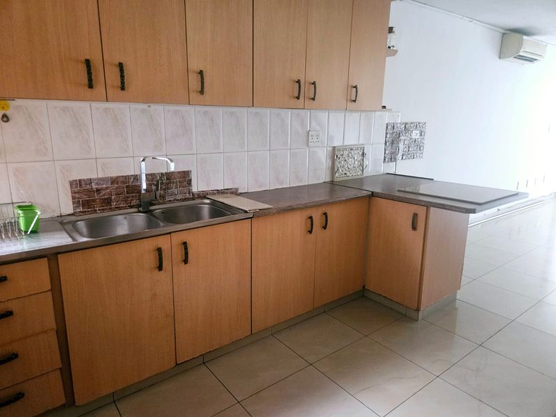 A functional kitchen featuring light wood-look laminate cabinetry, tiled backsplash, double stainless steel sink, and tiled flooring with an integrated air conditioning unit.