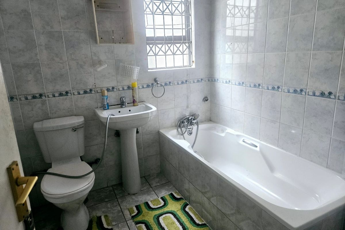 A functional bathroom featuring a pedestal basin, standard toilet, and bathtub with full-height wall tiling and security bars on the window.