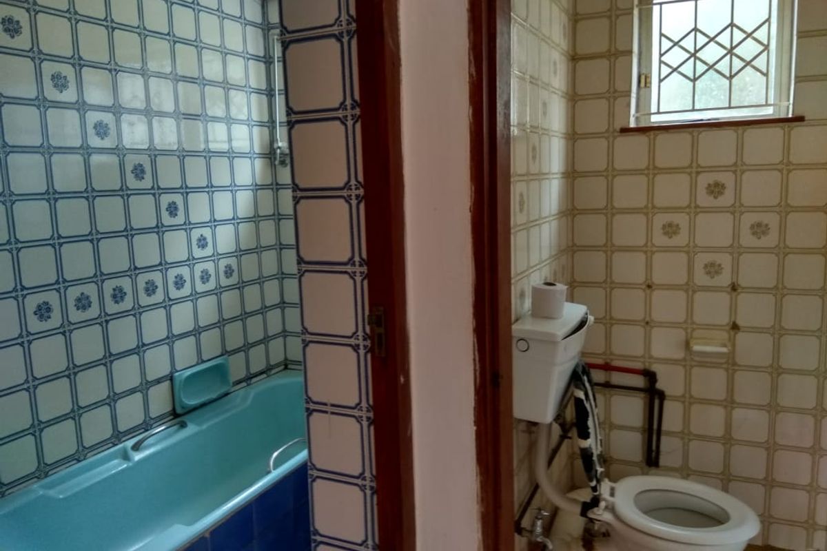 A dated bathroom featuring a built-in bathtub and separate toilet area with original ceramic wall tiling and a small window with security bars.