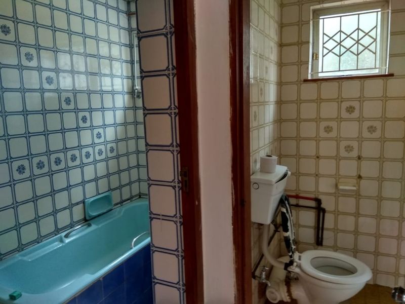 A dated bathroom featuring a built-in bathtub and separate toilet area with original ceramic wall tiling and a small window with security bars.