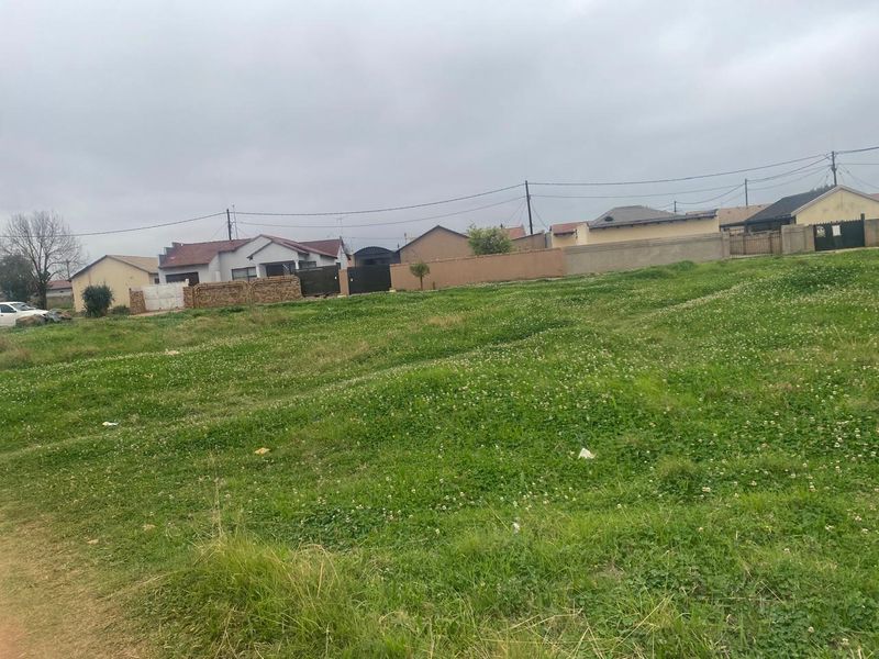 A vacant residential plot featuring a grass-covered surface with surrounding boundary walls in a developed neighborhood setting.
