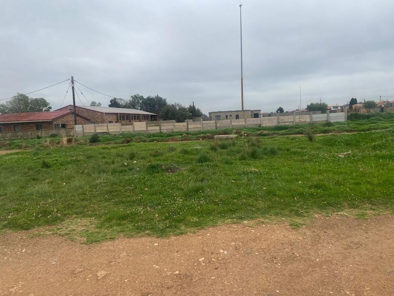 A vacant residential plot featuring grass coverage, enclosed by a precast concrete boundary wall with nearby utility infrastructure.