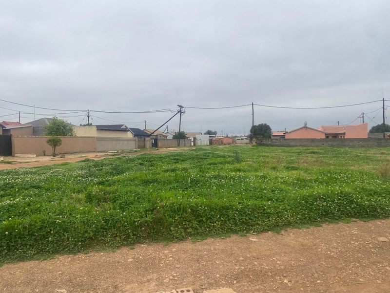 A vacant residential plot of land featuring grass cover, perimeter walls, and nearby utility infrastructure in a suburban setting.