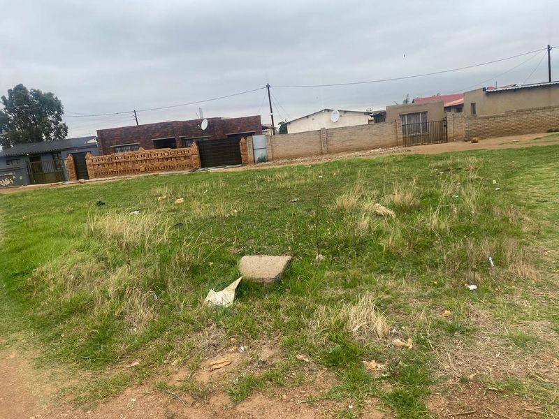 A vacant residential plot featuring grass cover, enclosed by perimeter walls and fencing in a developed neighborhood setting.