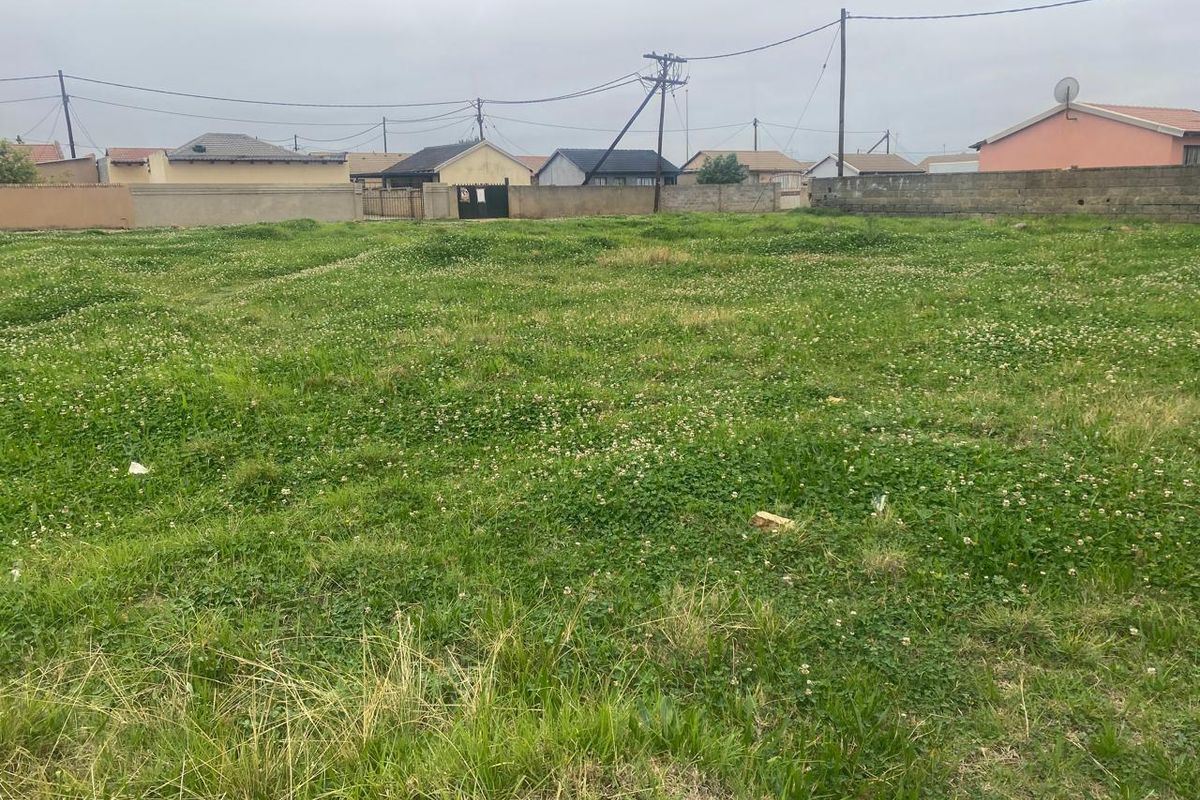 A vacant residential plot of land featuring grass coverage and perimeter boundary walls, situated in a developed neighborhood setting.