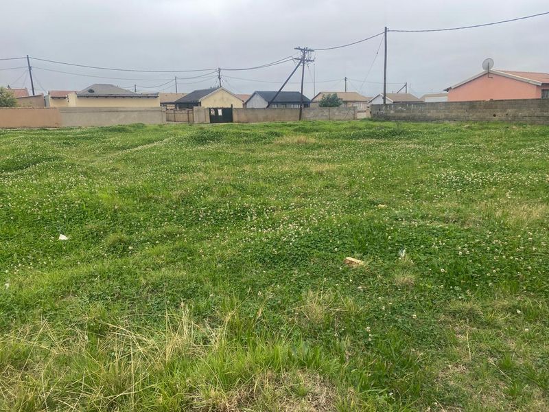 A vacant residential plot of land featuring grass coverage and perimeter boundary walls, situated in a developed neighborhood setting.