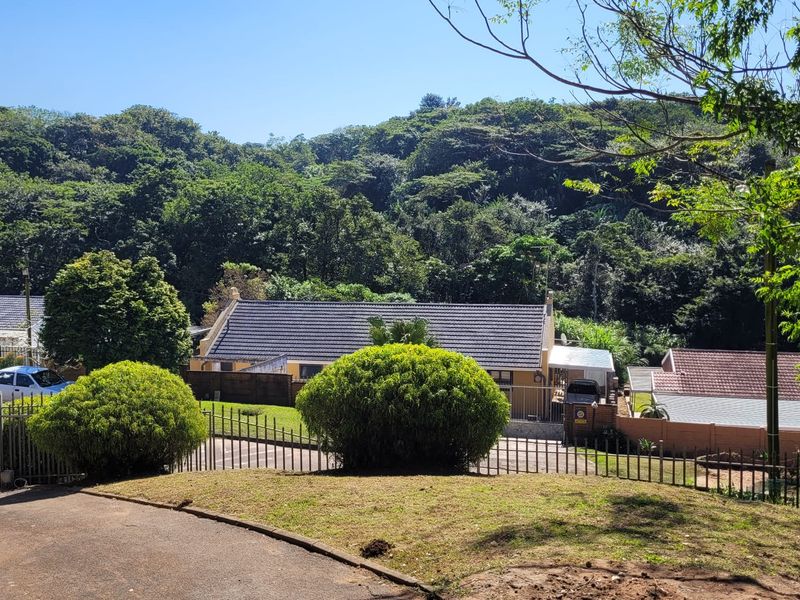 A view of the front exterior of the property featuring a tiled roof, paved driveway, and established garden set against a lush green backdrop.
