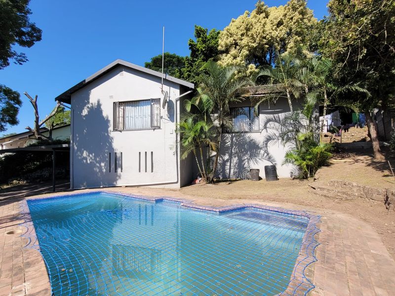 Outdoor pool area featuring a swimming pool with safety net, surrounded by brick paving and established garden landscaping.