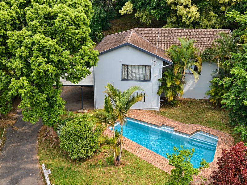 Aerial view of a residential property featuring a swimming pool with brick coping, a tiled roof, and a paved driveway surrounded by mature trees.