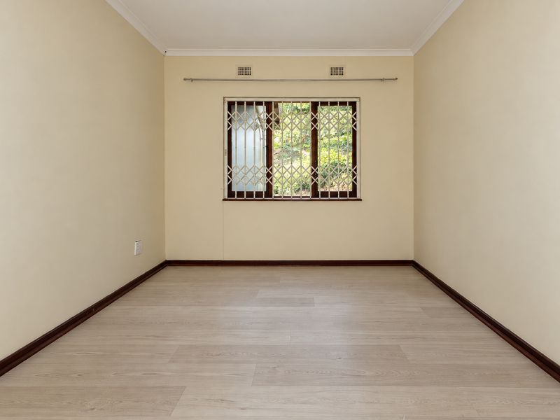 A clean, empty bedroom featuring light-toned laminate flooring, neutral walls, and a window fitted with a security gate.