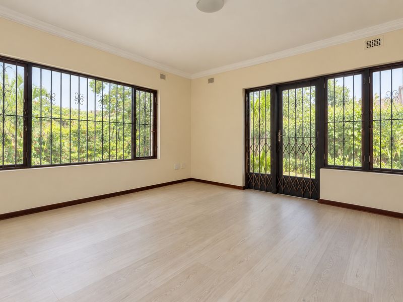A bright, empty living room featuring light-toned laminate flooring, large windows with security bars, and a secure access door leading to the garden.