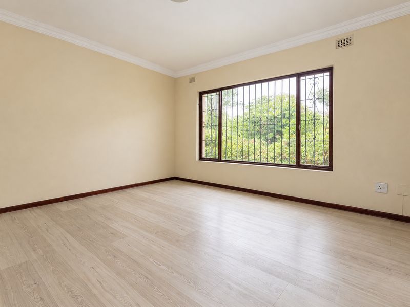 A bright, empty bedroom featuring light-toned laminate flooring, neutral walls, and a large window fitted with security bars.