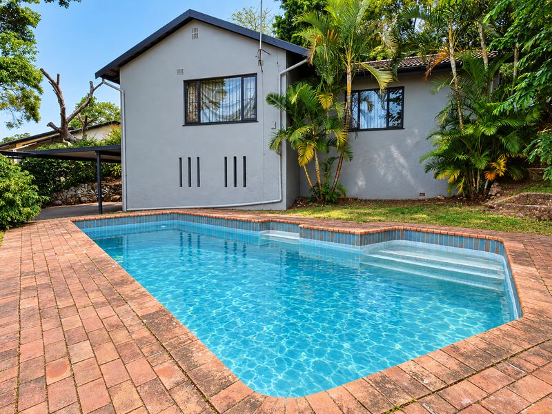 A well-maintained swimming pool area featuring brick paving, integrated pool steps, and a view of the rear building elevation with a carport.