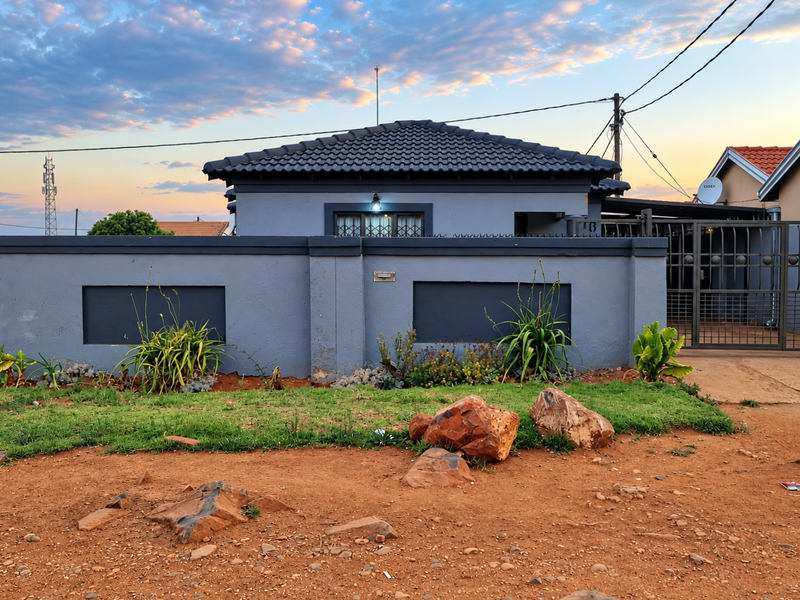 Front view of a residential property featuring a grey perimeter wall, metal security gate, and a dark tiled roof at sunset.