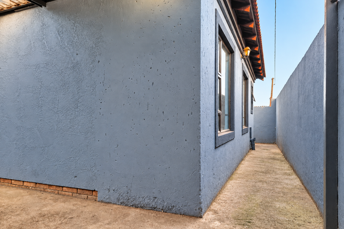 A side exterior view of the property featuring a concrete pathway, plastered walls, and exterior lighting along the building perimeter.
