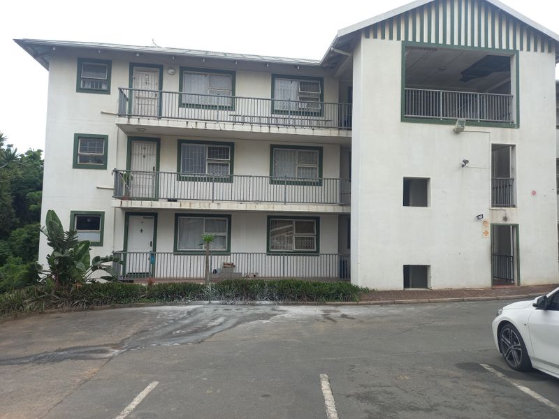 Exterior view of a multi-story apartment building featuring balconies with metal railings, security window bars, and an adjacent asphalt parking area.