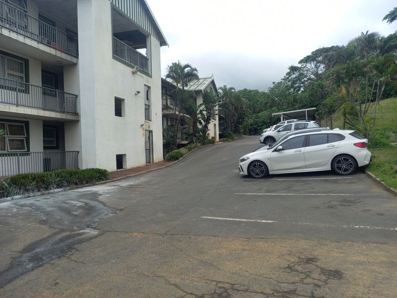 Exterior view of an apartment complex featuring a multi-story facade with balcony railings and an asphalt parking area with marked bays.