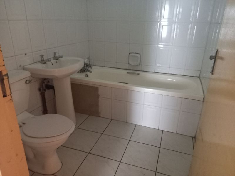 A functional bathroom featuring a pedestal basin, bathtub, and toilet with white ceramic wall and floor tiles, requiring some cosmetic updates to the paneling.