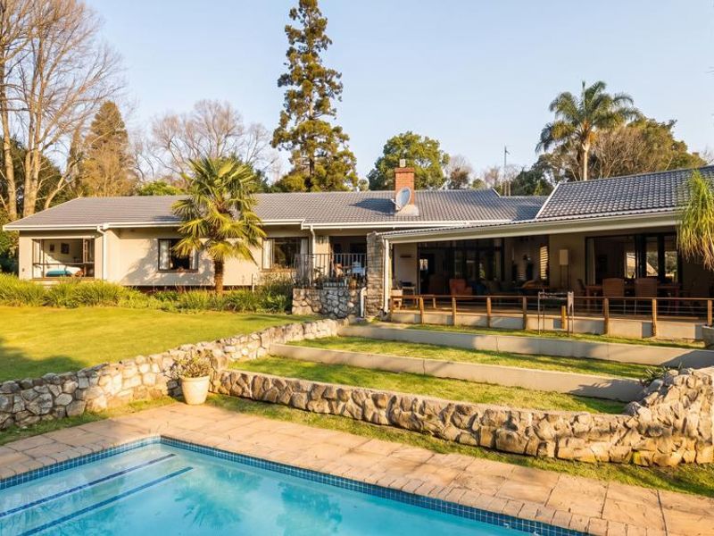 Spacious rear exterior view featuring a well-maintained swimming pool, stone retaining walls, a covered patio area, and a large landscaped garden.