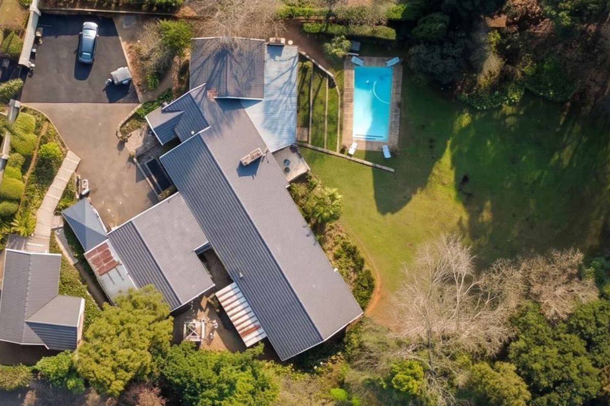 Aerial view of the property featuring a large garden, swimming pool, paved driveway, and a multi-section metal roof structure.