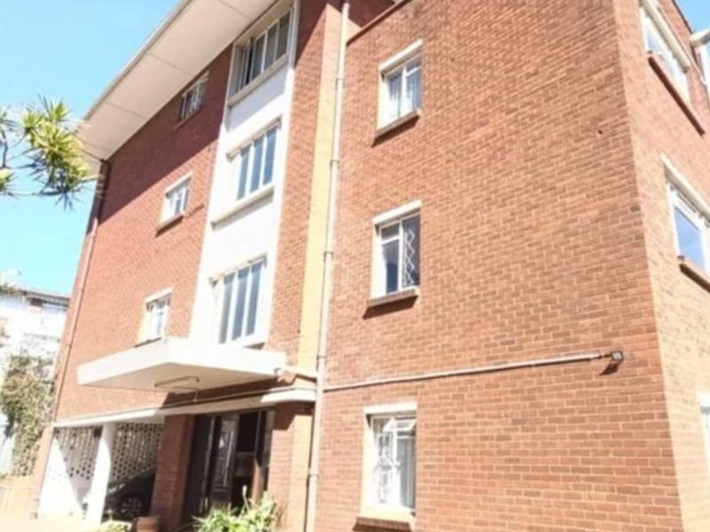 A multi-story apartment building exterior featuring a classic red brick facade, covered entrance area, and multiple window placements.