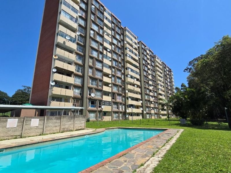 Exterior view of a multi-story apartment building featuring a communal swimming pool, paved surround, and landscaped garden area.