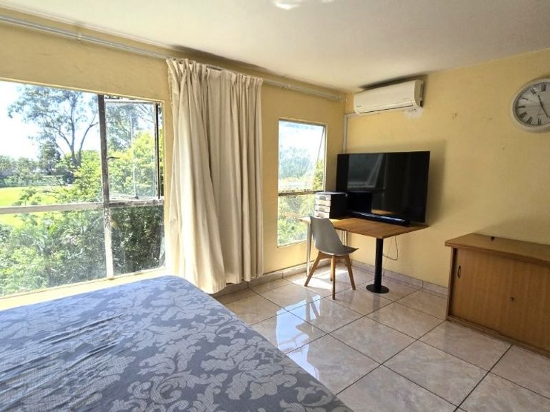 A bright bedroom featuring large windows with garden views, tiled flooring, and a wall-mounted air conditioning unit.