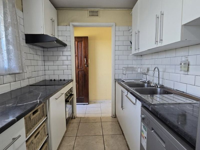 A functional galley-style kitchen featuring white cabinetry, subway tile backsplash, dark laminate countertops, and integrated cooking appliances with tiled flooring.