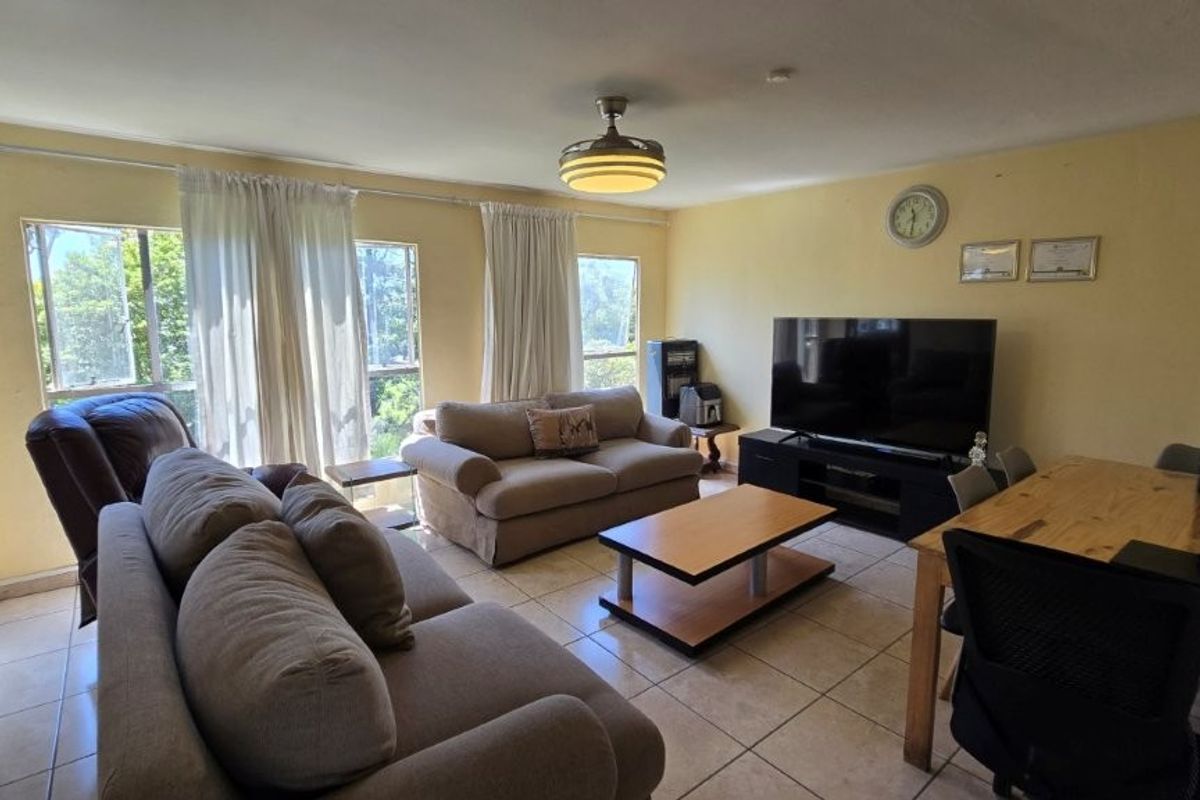A spacious living area featuring tiled flooring, a ceiling fan, and large windows that provide ample natural light.