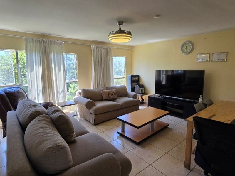 A spacious living area featuring tiled flooring, a ceiling fan, and large windows that provide ample natural light.