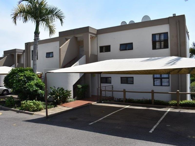 Exterior view of a townhouse complex featuring a paved parking area with covered carports and established landscaping.